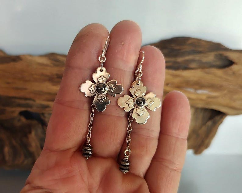 Pode incluir: Brincos de flor prateados com uma pedra escura no centro e detalhes de contas penduradas. Os brincos t&ecirc;m a forma de uma flor de quatro p&eacute;talas. Os brincos est&atilde;o pendurados em um gancho prateado.