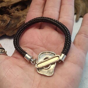 K&ouml;nnte beinhalten: Ein schwarzes, geflochtenes Lederarmband mit einem silbernen und goldenen Verschluss. Der Verschluss ist ein einzigartiges Design mit einer strukturierten Oberfl&auml;che.