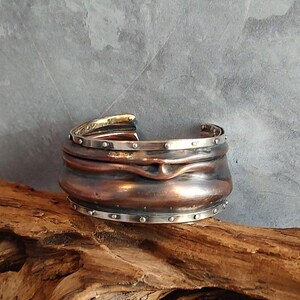 May include: A wide copper cuff bracelet with a hammered finish and a silver band with small silver dots around the edge.