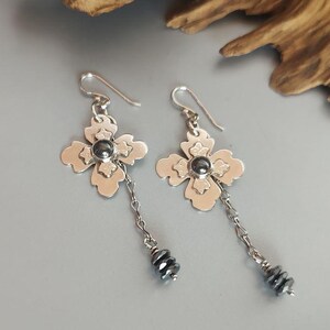 Pode incluir: Um par de brincos de prata com um design floral. Cada brinco apresenta uma flor de quatro p&eacute;talas com uma pedra central cinza escuro e uma corrente pendurada com pequenas contas cinza escuro. Os brincos s&atilde;o exibidos em uma superf&iacute;cie cinza claro.