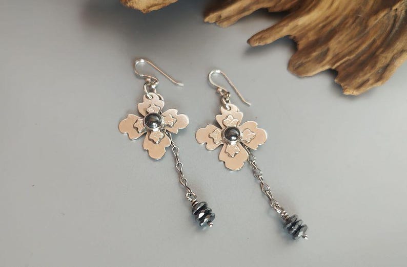 Pode incluir: Um par de brincos de prata com um design floral. Cada brinco apresenta uma flor de quatro p&eacute;talas com uma pedra central cinza escuro e uma corrente pendurada com pequenas contas cinza escuro. Os brincos s&atilde;o exibidos em uma superf&iacute;cie cinza claro.