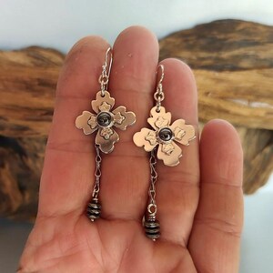 Pode incluir: Um par de brincos prateados. Cada brinco apresenta um design em forma de flor com uma pedra escura no centro e uma corrente pendurada que termina em um grupo de pequenas contas. Os brincos s&atilde;o presos por um gancho prateado.