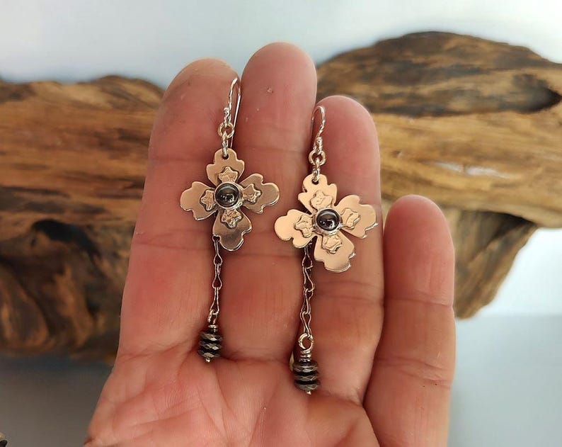 Pode incluir: Um par de brincos prateados. Cada brinco apresenta um design em forma de flor com uma pedra escura no centro e uma corrente pendurada que termina em um grupo de pequenas contas. Os brincos s&atilde;o presos por um gancho prateado.