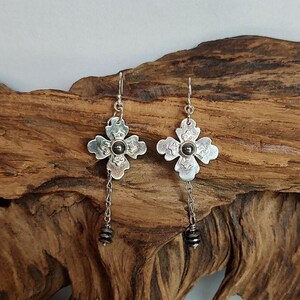 Pode incluir: Um par de brincos de flores prateados. Cada brinco apresenta um design de flor de quatro p&eacute;talas com uma pedra central cinza escuro. Uma corrente com pequenas contas cinza escuro pende de cada brinco, criando um visual delicado e elegante.