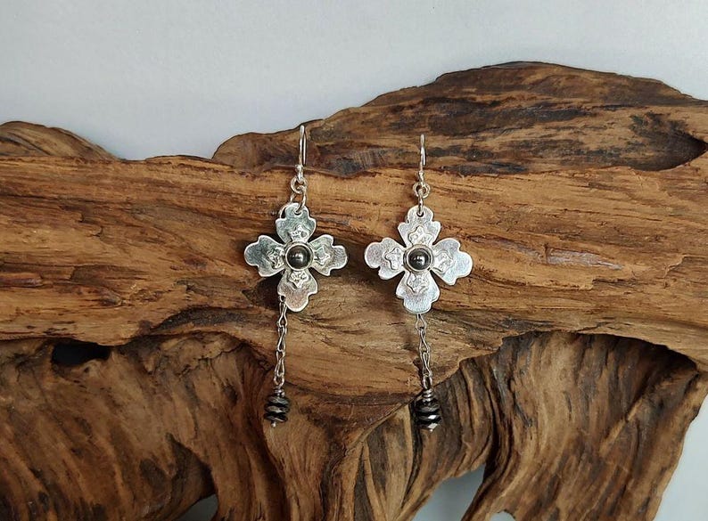 Pode incluir: Um par de brincos de flores prateados. Cada brinco apresenta um design de flor de quatro p&eacute;talas com uma pedra central cinza escuro. Uma corrente com pequenas contas cinza escuro pende de cada brinco, criando um visual delicado e elegante.
