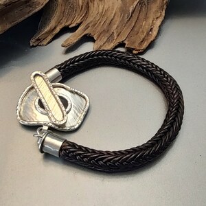 K&ouml;nnte beinhalten: Ein schwarzes geflochtenes Lederarmband mit einem silbernen und goldenen Verschluss. Der Verschluss ist ein einzigartiges Design mit einer strukturierten Oberfl&auml;che.
