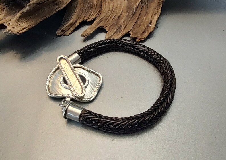 K&ouml;nnte beinhalten: Ein schwarzes geflochtenes Lederarmband mit einem silbernen und goldenen Verschluss. Der Verschluss ist ein einzigartiges Design mit einer strukturierten Oberfl&auml;che.