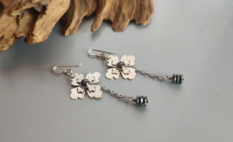 Pode incluir: Um par de brincos pendurados em tons de prata. Cada brinco apresenta um design de flor de quatro p&eacute;talas com um centro de contas cinza escuro. Uma corrente se estende da flor a um aglomerado de contas cinza escuro. Os brincos s&atilde;o exibidos em uma superf&iacute;cie cinza.