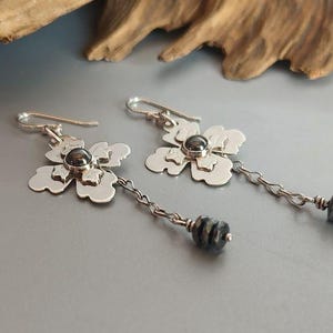 Pode incluir: Um par de brincos de prata com flores, com um centro de pedra cinza escuro e uma corrente pendurada com contas cinza escuro. Os brincos s&atilde;o exibidos em uma superf&iacute;cie cinza.
