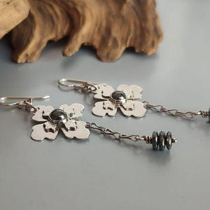 Pode incluir: Brincos de prata com flores, com um centro de contas cinza escuro e contas cinza escuro penduradas. Os brincos t&ecirc;m fecho de gancho e s&atilde;o exibidos em uma superf&iacute;cie cinza com um detalhe de madeira no fundo.