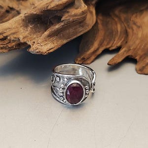 May include: A sterling silver ring with an oval, deep red gemstone. The ring has an ornate, textured design with small circular accents. The gemstone is the focal point, set in a detailed silver band. The ring is displayed on a light gray surface.