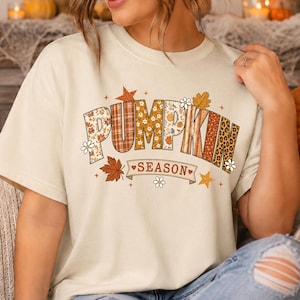May include: A cream-colored short-sleeve t-shirt featuring the text PUMPKIN SEASON in large block letters. The letters are filled with various autumn-themed patterns including plaid, floral, and leopard print, accented by small leaves, stars, and flowers.
