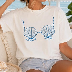 May include: A cream-colored short-sleeve t-shirt featuring a blue graphic of two seashells connected by wavy lines. The text "Ancha's bachelorette" is printed under the left shell and "Miami 2026" is printed under the right shell.