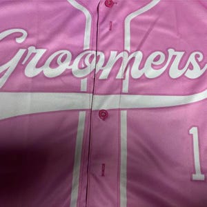 Puede incluir: Una camiseta de b&eacute;isbol rosa claro con botones que presenta la palabra Groomers en letras cursivas blancas grandes en el pecho. La prenda incluye un ribete vertical blanco a lo largo de la tapeta frontal y el n&uacute;mero 1 impreso en blanco a la derecha.