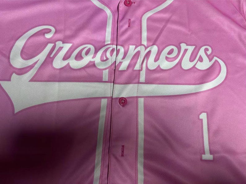 Puede incluir: Una camiseta de b&eacute;isbol rosa claro con botones que presenta la palabra Groomers en letras cursivas blancas grandes en el pecho. La prenda incluye un ribete vertical blanco a lo largo de la tapeta frontal y el n&uacute;mero 1 impreso en blanco a la derecha.