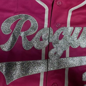 Puede incluir: Una camiseta de b&eacute;isbol de color rosa brillante con la palabra Rogue en letras grandes de purpurina plateada en la parte delantera. La prenda incluye un detalle de ribete blanco vertical a lo largo de la tapeta y dos botones rosas visibles.