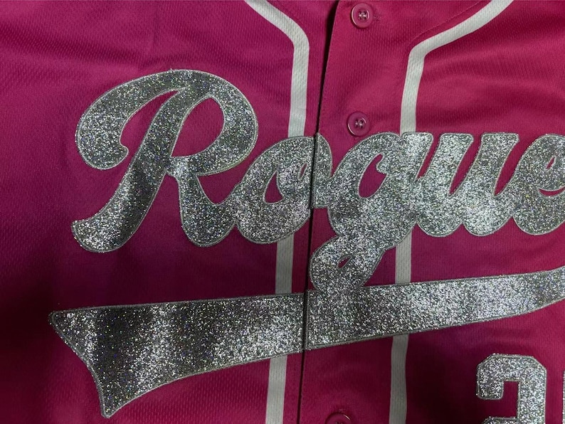Puede incluir: Una camiseta de b&eacute;isbol de color rosa brillante con la palabra Rogue en letras grandes de purpurina plateada en la parte delantera. La prenda incluye un detalle de ribete blanco vertical a lo largo de la tapeta y dos botones rosas visibles.