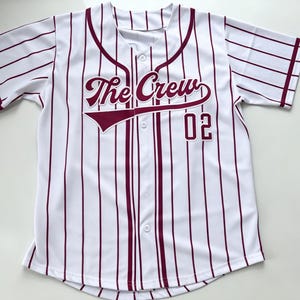 Puede incluir: Una camiseta de b&eacute;isbol blanca de manga corta con rayas verticales de color granate, cierre frontal de botones y el texto The Crew junto al n&uacute;mero 02 impreso en granate en el pecho, con ribetes granates en el cuello y los pu&ntilde;os.