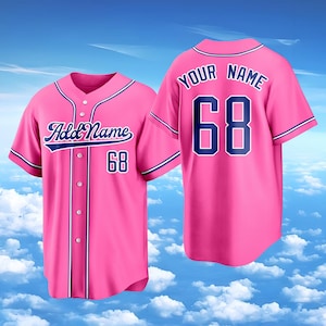 Puede incluir: Una camiseta de b&eacute;isbol de color rosa brillante con ribetes y letras en azul oscuro. La parte delantera muestra el texto Add Name y el n&uacute;mero 68. La parte trasera muestra el texto YOUR NAME sobre el n&uacute;mero 68 con cierre de botones.