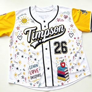 Puede incluir: Una camiseta de b&eacute;isbol blanca con mangas amarillas y ribetes negros. El frente presenta el texto Timpson, el n&uacute;mero 26 y las palabras Learn Love Inspire. Los gr&aacute;ficos incluyen un sol, corazones, estrellas y una pila de libros con una manzana.