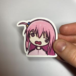 Puede incluir: Una pegatina dibujada a mano con un personaje de estilo anime con pelo rosa y una expresión de sorpresa. El personaje tiene ojos grandes y lleva un atuendo rosa. La pegatina está delineada en negro y tiene un borde blanco.