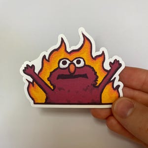 Könnte beinhalten: Ein Aufkleber mit einer Cartoonfigur, die von Flammen umgeben ist. Die Figur ist tiefrot mit großen weißen Augen und orangefarbenen Flammen. Die Arme der Figur sind erhoben, und der Aufkleber hat einen weißen Rand.