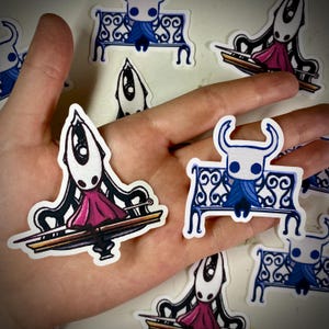 Puede incluir: Colección de pegatinas con un personaje con cabeza blanca, detalles negros y vestido rosa. El personaje está sentado en un banco azul con detalles ornamentados. Las pegatinas están en una mano.