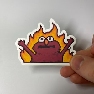 Op de afbeelding: Een sticker met een stripfiguur, Elmo, omringd door vlammen. Het personage is dieprood met witte ogen en oranje vlammen. De sticker heeft een witte rand en wordt door een hand vastgehouden.