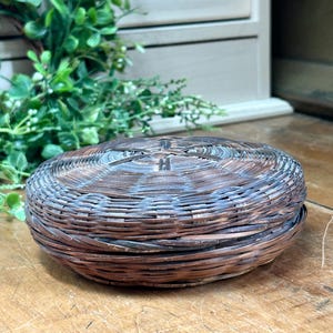 May include: A round, woven basket with a lid. The basket is made of brown and gray-blue colored reeds. The lid features a cross-like design. The basket sits on a wooden surface, with greenery in the background.