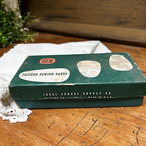 May include: A rectangular dark green cardboard box labeled Ideal Pricked Sewing Cards. The lid features white graphic shapes with illustrations of stitched patterns and the text Ideal School Supply Co. Chicago 20, Illinois, Made in U.S.A.