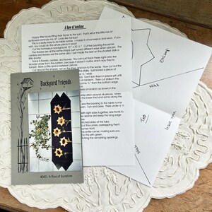 May include: A collection of craft supplies, including a pattern for a table runner with sunflower designs, instructions, and diagrams. The pattern features the text "A Row of Sunshine" and "Backyard Friends."