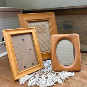 May include: A collection of three wooden picture frames in varying sizes and shapes. Two frames are rectangular with flat edges, while the third frame features a rounded, oval-shaped inner opening. Each frame contains a plain cardboard backing.