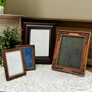 May include: Three rectangular picture frames of varying sizes. One frame is dark brown with a smooth finish, one is a smaller brown frame with a textured surface, and one is a light brown frame with ornate carved corner details.