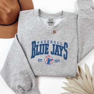 May include: A grey sweatshirt with the text "BASEBALL BLUE JAYS" in blue block letters. The sweatshirt also features a baseball graphic with the text "EST. 1977". A brown hat is visible in the background.