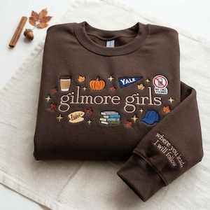 Peut inclure: Un sweat-shirt à col rond marron foncé avec des broderies représentant une tasse de café, une citrouille, un fanion Yale, un panneau interdiction de téléphones, une enseigne Luke's, des livres et une casquette bleue. Le texte "gilmore girls" est centré, avec "where you lead, I will follow" sur la manche.