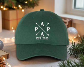 May include: A dark green baseball cap with the letters "P A P A" in white, crossed with arrows, and the text "EST. 2025". The cap is made of a soft material and has a curved brim. The cap is a classic style.