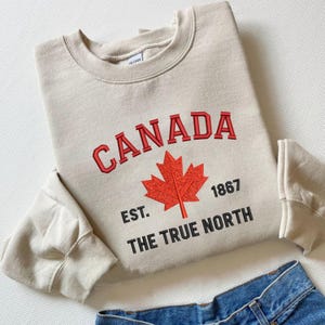 Op de afbeelding: Een beige sweatshirt met het woord "CANADA" in rode geborduurde letters, een rood esdoornblad en de tekst "EST. 1867 THE TRUE NORTH". De sweatshirt is opgevouwen, met denim jeans zichtbaar aan de onderkant.