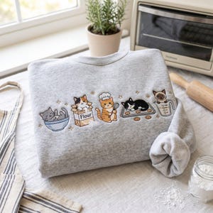 May include: A heather gray crewneck sweatshirt featuring an embroidered design of five cats baking. The cats are shown in a bowl, a flour bag, wearing a chef hat with a whisk, on a cookie sheet, and inside a measuring cup with small stars.