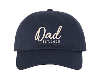 May include: Navy blue baseball cap with the word "Dad" in a cursive font, and the text "EST. 2025" below it. The cap has a curved brim and a classic six-panel design, suitable for casual wear.
