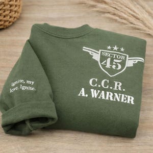 May include: An olive green sweatshirt with white text and graphics. The design includes the words "Sector 45", "C.C.R. A. Warner", and the phrase "Ignite, my love. Ignite." The sweatshirt is folded on a wooden surface.