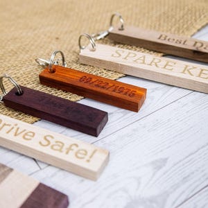 May include: Five wooden keychains with different engraved text and colors. The keychains are made of different types of wood, including light brown, dark brown, and purple. The text on the keychains includes "Spare Key", "Best Dad", "09/22/1975", "Drive Safe!" and "The Crew".