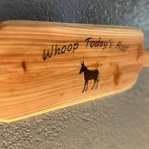 May include: Wooden cutting board with a handle, featuring the text "Whoop Today's Ass!" and a silhouette of a donkey. The board is made of light-colored wood with visible grain patterns, perfect for kitchen decor.