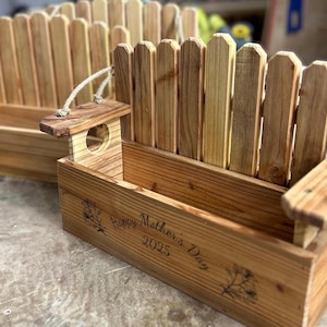 Puede incluir: Dos jardineras de madera con diseño de valla. Cada jardinera tiene una caja rectangular con respaldo en forma de valla y un pequeño asa de madera con un agujero. El texto "Happy Mother's Day 2025" está grabado en el frente.