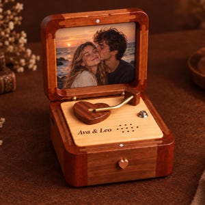 Puede incluir: Una caja de música de madera con tapa abatible que muestra una foto de una pareja al atardecer. La base incluye un brazo de tocadiscos en forma de corazón, un pequeño botón plateado y un panel de madera clara grabado con Ava & Leo.