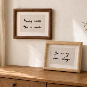Pode incluir: Dois porta-retratos retangulares com inserções de tecido claro e texto bordado em preto. A moldura superior é de madeira castanha escura com o texto "Family makes this a home." A moldura inferior é de madeira clara com "You are my home, always."