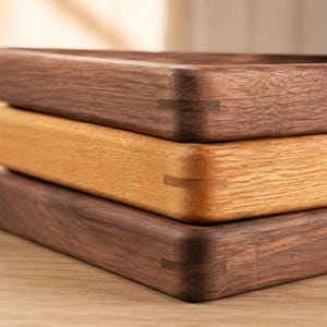 May include: A stack of three rectangular wooden trays with rounded corners. The top and bottom trays are made of dark brown walnut wood, while the middle tray is made of light tan oak wood. Each tray features visible spline joinery details.