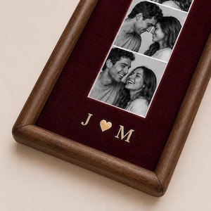 May include: A rectangular wooden picture frame featuring a deep burgundy velvet backing. The frame displays two black and white photographs of a couple. Below the photos, the gold-colored letters J and M are separated by a gold heart symbol.