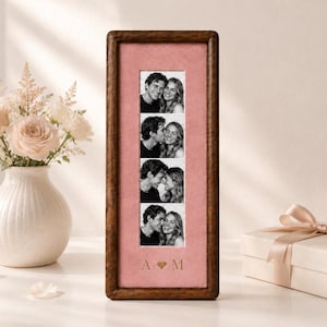 May include: A tall, rectangular wooden picture frame with a dark brown finish, featuring a vertical strip of four black and white photographs of a couple. The photos are set against a dusty pink mat with the gold-colored letters A and M separated by a heart.