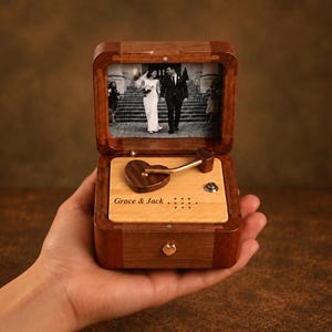 Op de afbeelding: Een rechthoekige houten muziekdoos met een donkerbruine afwerking. De binnenkant van het deksel toont een zwart-witfoto van een koppel. De basis heeft een licht houten paneel met de tekst Grace & Jack, een hartvormige arm en een metalen knop.