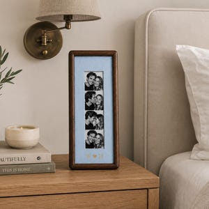 May include: A vertical rectangular dark wood photo frame with a light blue mat board. The frame displays a strip of four black and white photographs of a couple. Below the photos, the letters W and H are separated by a small heart symbol.